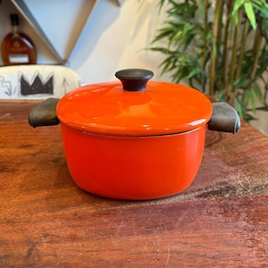 May include: Orange enamel cast iron pot with a black lid and black handles. The pot is sitting on a wooden surface.