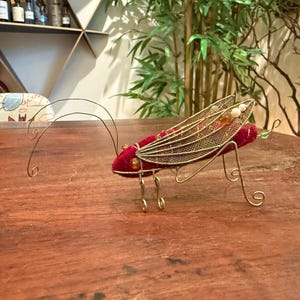 以下が含まれることがあります： 装飾的なバッタの置物。 体は深紅色で、金色のメッシュの羽と繊細なワイヤーの脚と触角が付いています。 この置物は、小さくて丸いオレンジと白のビーズで装飾されています。