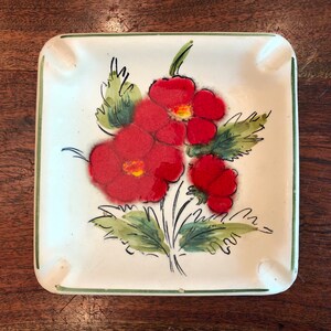 May include: Square ceramic ashtray with a green border and a floral design. The design features three red flowers with yellow centers and green leaves.