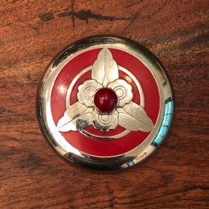 Könnte beinhalten: Ein runder silberner Deckel mit einer roten Mitte und einem silbernen Blumendesign mit einem roten Juwel in der Mitte.
