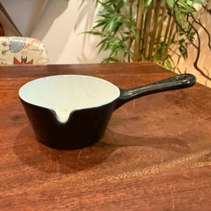 Puede incluir: Un cazo pequeño y redondo de hierro fundido negro con un revestimiento interior de esmalte blanco. El cazo cuenta con un mango largo y un pequeño pico vertedor en el borde, diseñado para facilitar el vertido de líquidos.