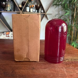 May include: A vintage, deep red glass bottle with a rounded top and a ribbed base sits next to a plain, brown cardboard box. The bottle has a smooth, glossy finish. The box is rectangular and shows signs of wear.