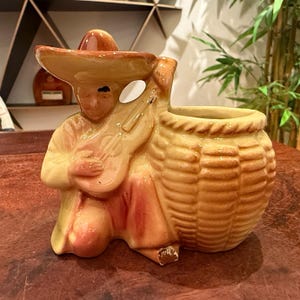 May include: A glazed ceramic figurine of a person wearing a sombrero and playing a guitar, attached to a textured, basket-shaped planter. The piece features warm tan, brown, and cream tones with a glossy finish and a small area of paint loss.