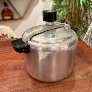 May include: A vintage silver pressure cooker with a black handle and knob. The cooker has a round body and a lid with a pressure release valve. The cooker is sitting on a wooden surface.