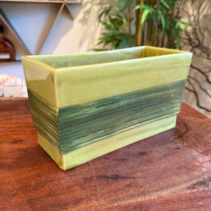 Op de afbeelding: Een rechthoekige, lichtgroene keramische plantenbak met een gestreepte, donkergroene band in het midden. De plantenbak heeft een glanzende afwerking en is ontworpen voor binnen of buiten. De plantenbak is ongeveer 25 cm lang.