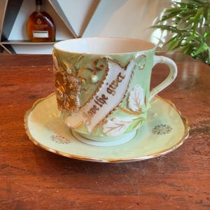 Puede incluir: Una taza de té y un platillo verdes con ribetes dorados. La taza de té tiene una cinta blanca con las palabras "Love the giver" en letras doradas. El platillo tiene un diseño de copo de nieve dorado.