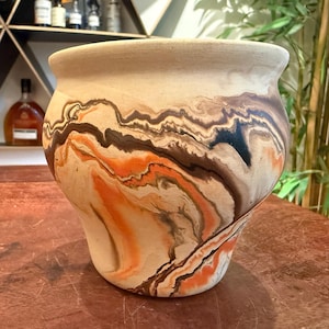 May include: A rounded ceramic planter featuring a marbled surface design with swirling patterns of cream, burnt orange, dark brown, and black. The pot has a wide, flared rim and a smooth, matte finish, showcasing a fluid, organic color blend.