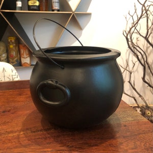 May include: A black cauldron with a handle and a ring on the side. The cauldron is sitting on a wooden surface. The cauldron is a prop for Halloween or other themed events.