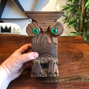 Pode incluir: Uma escultura de coruja de madeira com olhos de vidro verde. A coruja é esculpida com detalhes intrincados e tem um acabamento marrom rústico.