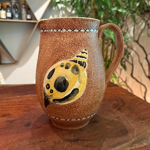 May include: A brown ceramic pitcher with a textured, speckled surface. The body features a painted yellow and black seashell motif on the front. A decorative band with a white cross-stitch pattern encircles the top and bottom of the vessel.