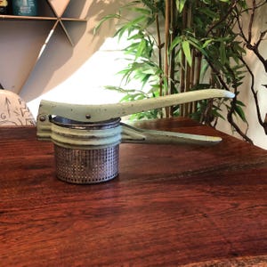 May include: A vintage green metal potato masher with a perforated metal base. The masher is on a wooden table.