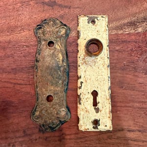 May include: Two antique door hardware pieces. One is a decorative, ornate backplate with a tarnished gold finish. The other is a rectangular plate with a keyhole and a circular knob opening, painted in a distressed cream color.