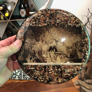 Puede incluir: Un posavasos redondo de resina transparente con una fotografía vintage de las cuevas de Carlsbad, Nuevo México. La foto muestra la entrada a la Sala Grande, con el texto "Carlsbad Caverns" y "Entrance to the Big Room" impreso en la foto. El posavasos está decorado con pequeñas piedras de colores alrededor del borde.