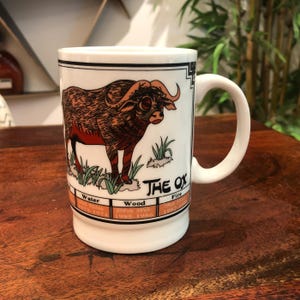 Puede incluir: Taza de cerámica blanca con una ilustración marrón y negra de un buey. La taza tiene un borde negro alrededor de la ilustración y las palabras "The Ox" en texto negro. La taza también tiene un gráfico con los elementos del zodíaco chino de agua, madera y fuego, junto con los años asociados a cada elemento.