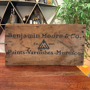 May include: A rectangular wooden crate with a weathered brown finish. The front panel features black printed text reading Benjamin Moore & Co., Est. 1883, and Paints-Varnishes-Muresco, centered around a triangular logo with the letters M and A.