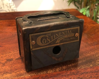 Op de afbeelding: Een vintage metalen spaarpot met een messing plaatje met de tekst "The Continental Shannon & Neville Winona, Minn."