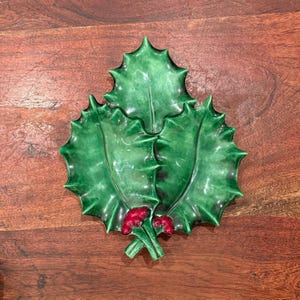 Puede incluir: Un plato de cerámica con forma de hoja de acebo con tres hojas verdes y bayas rojas. El plato es una decoración festiva, perfecta para la temporada navideña. El plato está colocado sobre una superficie de madera.