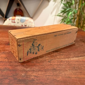 May include: A rectangular wooden box with a lid, featuring a printed graphic of a castle in blue and red ink. The side of the box is stamped with the text TWO POUNDS NET WEIGHT and FAILY CHEESE COMPANY - GREEN BAY, WISCONSIN in black ink.