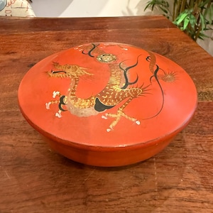 Könnte beinhalten: Runde, rote dekorative Box mit Deckel. Der Deckel zeigt eine detaillierte Illustration eines goldenen Drachen mit schwarzen und weißen Akzenten. Die Box besteht aus einem festen Material und steht auf einer Holzoberfläche.