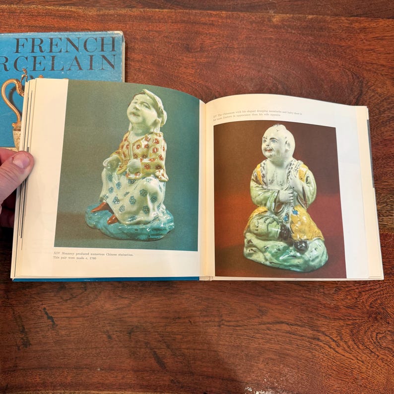 May include: Open book with images of antique porcelain figurines. The figurines are seated, with detailed clothing and facial features. The book's cover reads "FRENCH PORCELAIN" in blue and white.