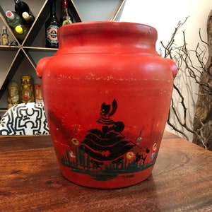 May include: A red ceramic jar with a black silhouette of a woman in a dress and flowers painted on the front.