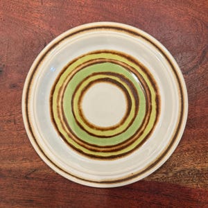 May include: A round ceramic plate featuring a series of concentric circles in shades of light green and dark brown against an off-white background. The plate has a smooth, glazed finish with a raised rim and a simple, circular design pattern.