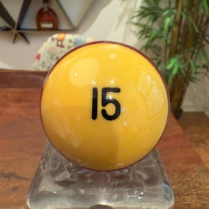 May include: A yellow billiard ball with the number 15 in black. The ball has a glossy finish and a reddish-brown band around the edge. It is resting on a clear glass base.