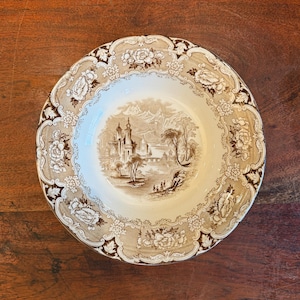 Może przedstawiać: Antyczna ceramiczna zupa z brązowo-białym wykończeniem, z kwiatowym obramowaniem i sceną zamku w górzystym krajobrazie.