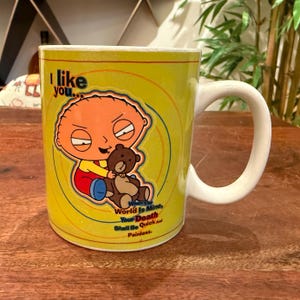 May include: A yellow ceramic mug featuring a cartoon character holding a brown teddy bear. The text reads: I like you... When The World Is Mine, Your Death Shall Be Quick And Painless. The mug has a white handle and a circular graphic design.