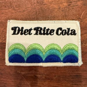 May include: A rectangular embroidered patch with an off-white background. The text Diet Rite Cola is stitched in black script across the top. The bottom half features a repeating pattern of overlapping scalloped arches in lime, teal, and blue.