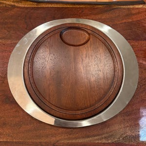 Op de afbeelding: Een cirkelvormige serveerplank met een donkerbruin houten middenstuk met een gladde afwerking en een verzonken ovale inkeping nabij de bovenrand. Het houten middenstuk is omgeven door een brede, gepolijste zilverkleurige metalen rand met een reflecterend oppervlak.
