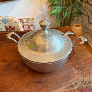 May include: A silver-colored metal serving dish with a lid and two handles. The dish has a round shape with a decorative lid and a small knob on top. The handles are curved and attached to the sides of the dish.