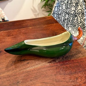 Könnte beinhalten: Ein grüner, chili-förmiger Keramik-Pflanztopf mit cremefarbenem Innenraum und orangefarbenem Stiel. Der Pflanztopf ist glänzend und steht auf einer Holzoberfläche. Das Design ist ein einzigartiger Dekorationsartikel.