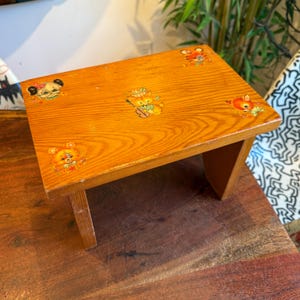 Puede incluir: Un pequeño taburete de madera vintage con un acabado marrón claro. La superficie superior presenta calcomanías de animales de dibujos animados coloridos, incluyendo un oso, un gato y un ciervo. El taburete tiene cuatro patas y probablemente se usa para niños.