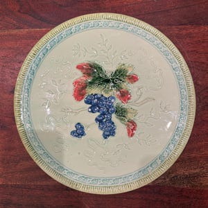 May include: A round ceramic plate with a cream-colored center featuring a raised, embossed design of a cluster of blue grapes and green and red leaves. The rim has a light blue patterned border and a textured, ribbed outer edge.