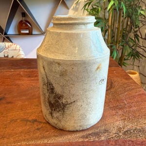 Puede incluir: Un tarro de gres antiguo, de color blanquecino, con un cuerpo cilíndrico y un cuello ligeramente ensanchado. El tarro tiene un aspecto rústico y envejecido con marcas de color marrón oscuro y una superficie texturizada. Probablemente se utiliza para almacenamiento o decoración.