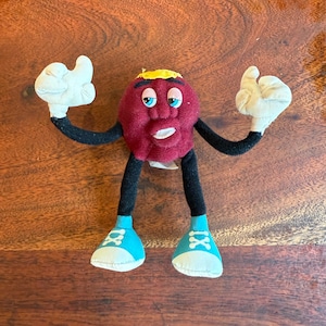 May include: A plush toy raisin character with a maroon body, black arms and legs, and white gloves. The character has blue eyes, a yellow band on its head, and is wearing blue trainers with white laces.