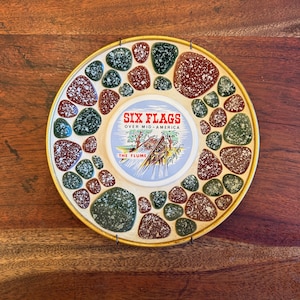 May include: A round decorative plate with a gold rim and a mosaic-style border of green and brown stones. The center features a circular design with the words "SIX FLAGS OVER MID-AMERICA" and an illustration of a water ride.