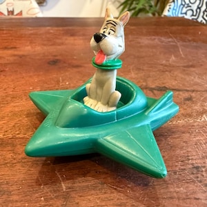 May include: A vintage toy featuring a cartoon dog figure with a red tongue and green collar, seated in a green star-shaped vehicle. The toy is made of plastic and has the word "Snoopy" printed on the side.