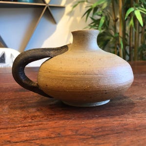 May include: A small, round, tan ceramic vessel with a speckled texture and a wide, low-profile body. It features a single, dark brown, curved handle attached to the side, leading up to a narrow, circular opening at the top of the piece.