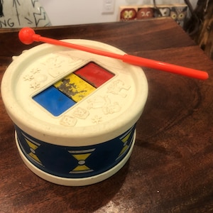 Könnte beinhalten: Eine weiße Spielzeugtrommel mit einem blauen, gelben und roten Design. Die Trommel hat einen roten und weißen Stock.