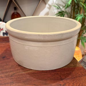 May include: A round, off-white ceramic crock with a smooth, glazed finish and a thick, reinforced rim. The vessel features a simple, cylindrical shape with a slightly wider top edge and a flat base, presenting a clean and functional design.