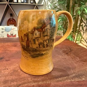 May include: A tan ceramic beer mug featuring a detailed illustration of a harbor scene with boats, buildings, and figures. The text 'Quay side Devon' is printed on the lower left side. The mug has a smooth, glossy finish and a large curved handle.