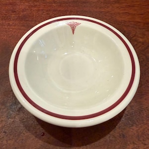 Peut inclure: Un bol rond en céramique blanc cassé avec un bord circulaire rouge foncé et un petit symbole de caducée rouge détaillé imprimé en haut au centre du bord intérieur. Le bol présente une finition lisse et brillante.