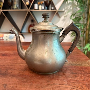 May include: Antique silver-colored teapot with a wooden handle and lid. The teapot has a spout and a decorative finial on the lid. The handle is dark brown and curved. The teapot is sitting on a wooden table.