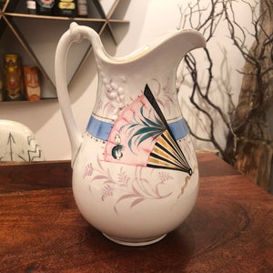 May include: White ceramic pitcher with a floral design and a pink and black fan painted on the side.
