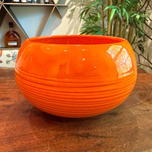 Puede incluir: Un cuenco de cerámica naranja vibrante con un acabado brillante. El cuenco tiene una forma redondeada con detalles horizontales acanalados alrededor del exterior. El interior es liso y el cuenco se asienta sobre una superficie de madera.