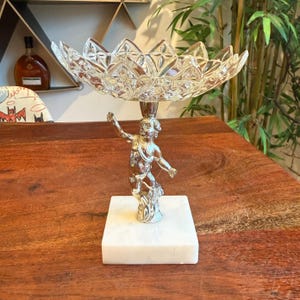 May include: A decorative centerpiece featuring a clear, cut-glass bowl supported by a silver-colored cherub figurine atop a white marble base. The bowl has a flower-like design. The piece is displayed on a wooden surface.