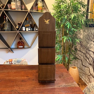 Puede incluir: Un portacartas alto de madera con tres compartimentos. La madera marrón oscuro tiene una veta visible. Un emblema de águila dorada está fijado en la parte superior. El portacartas está sobre una mesa de madera.