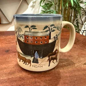 Puede incluir: Taza de cerámica con una ilustración pintada a mano del Arca de Noé. La taza presenta un cielo azul, un arca marrón y varios animales. La palabra "NOAH" está escrita en la parte delantera de la taza.
